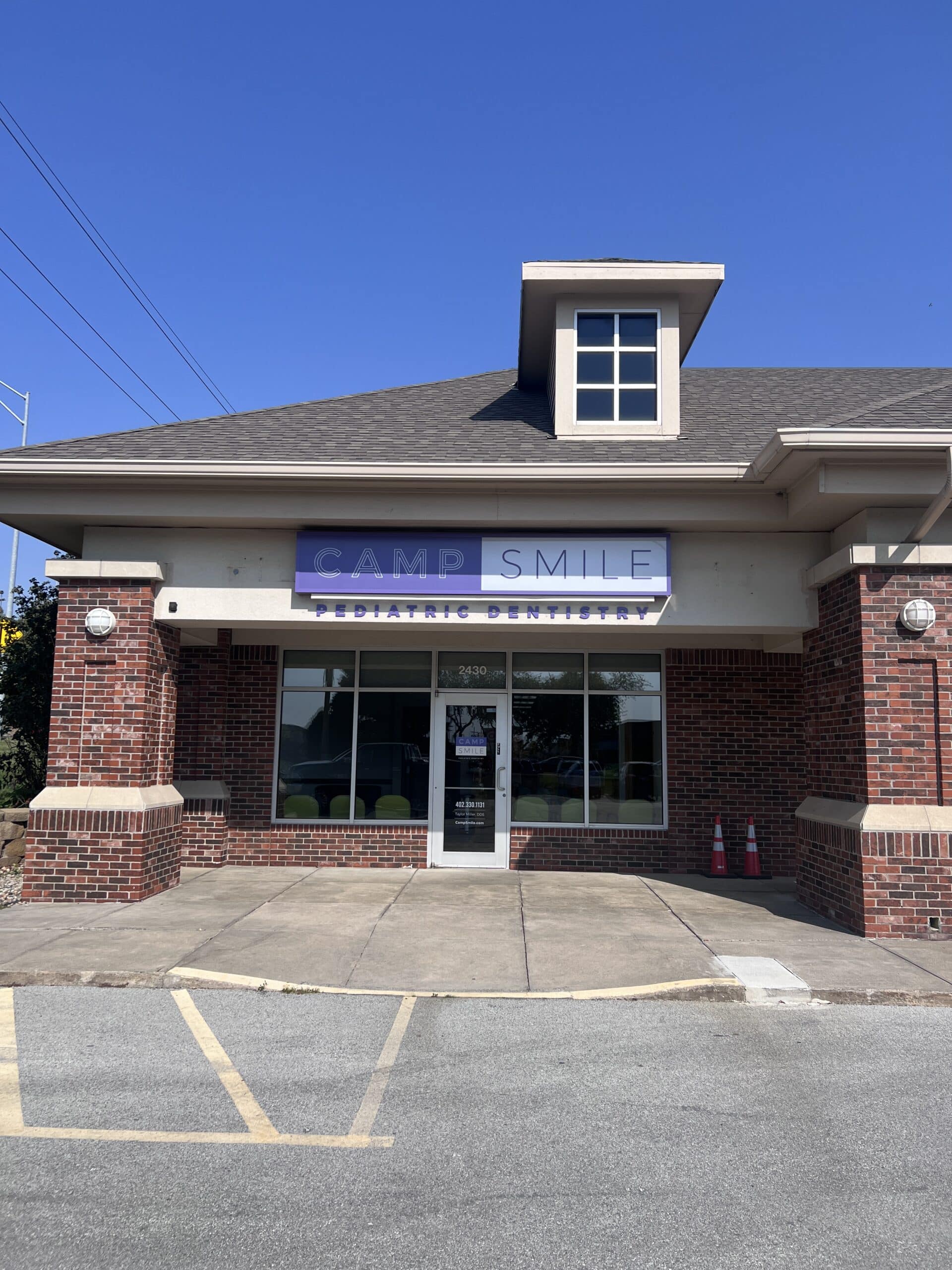 Brick building with "Camp Smile Pediatric Dentistry" sign, featuring large windows and a glass door. Two orange cones are placed near the entrance.