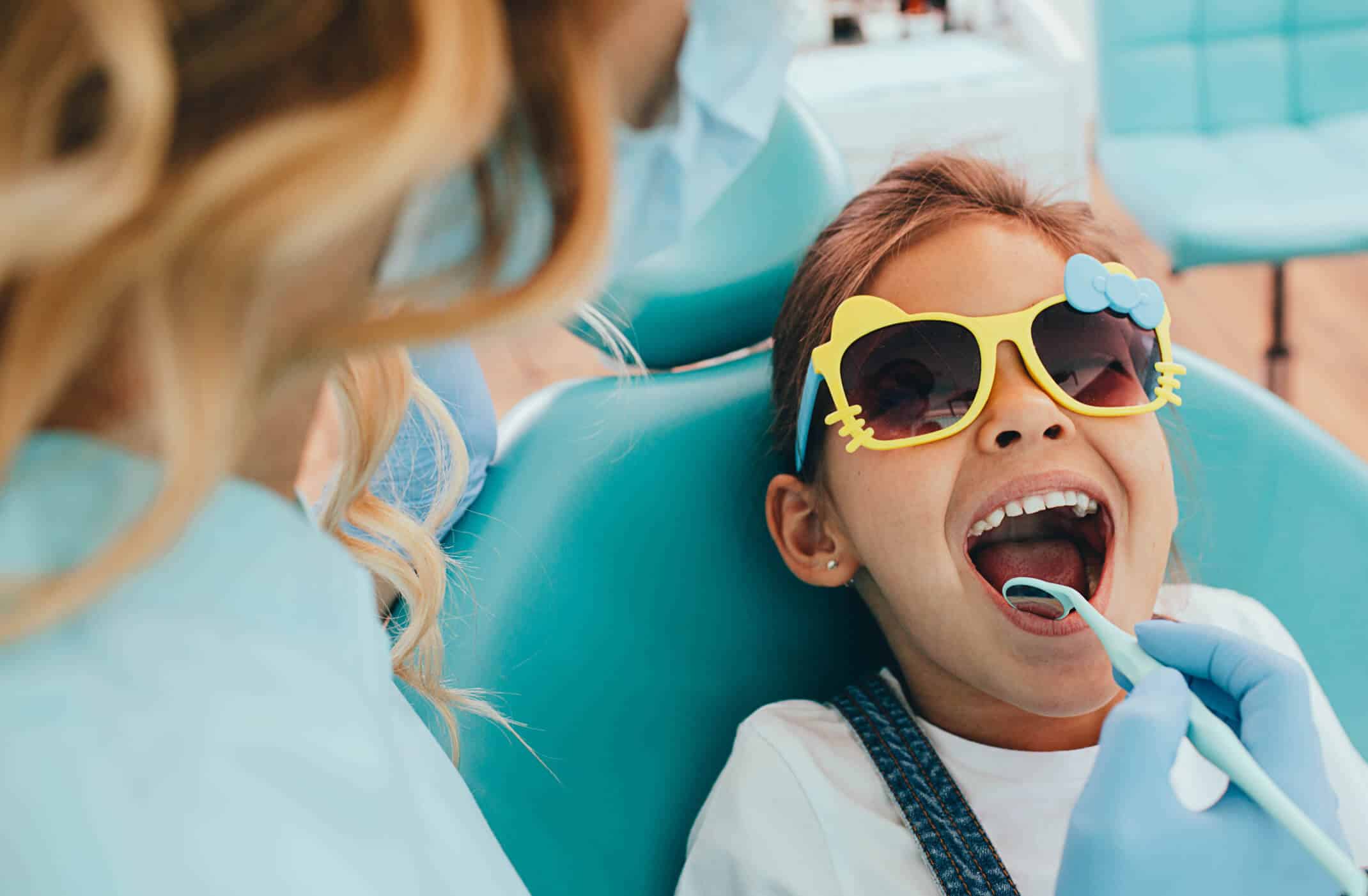 Girl wearing yellow and blue glasses in green dentist chair getting teeth cleaned.