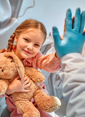 A child holding a teddy bear gives a high-five to a person wearing blue medical gloves in a warmly lit, clinical setting.