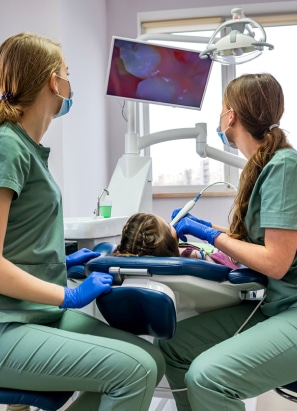 Two persons in green attire attend to a child patient in a dental clinic, with dental equipment and a screen displaying an image in the background.