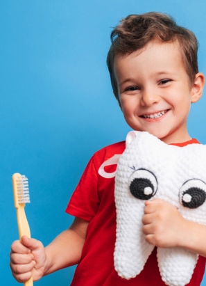 A child in a red shirt holds a toothbrush and a plush tooth with eyes, smiling against a blue background.