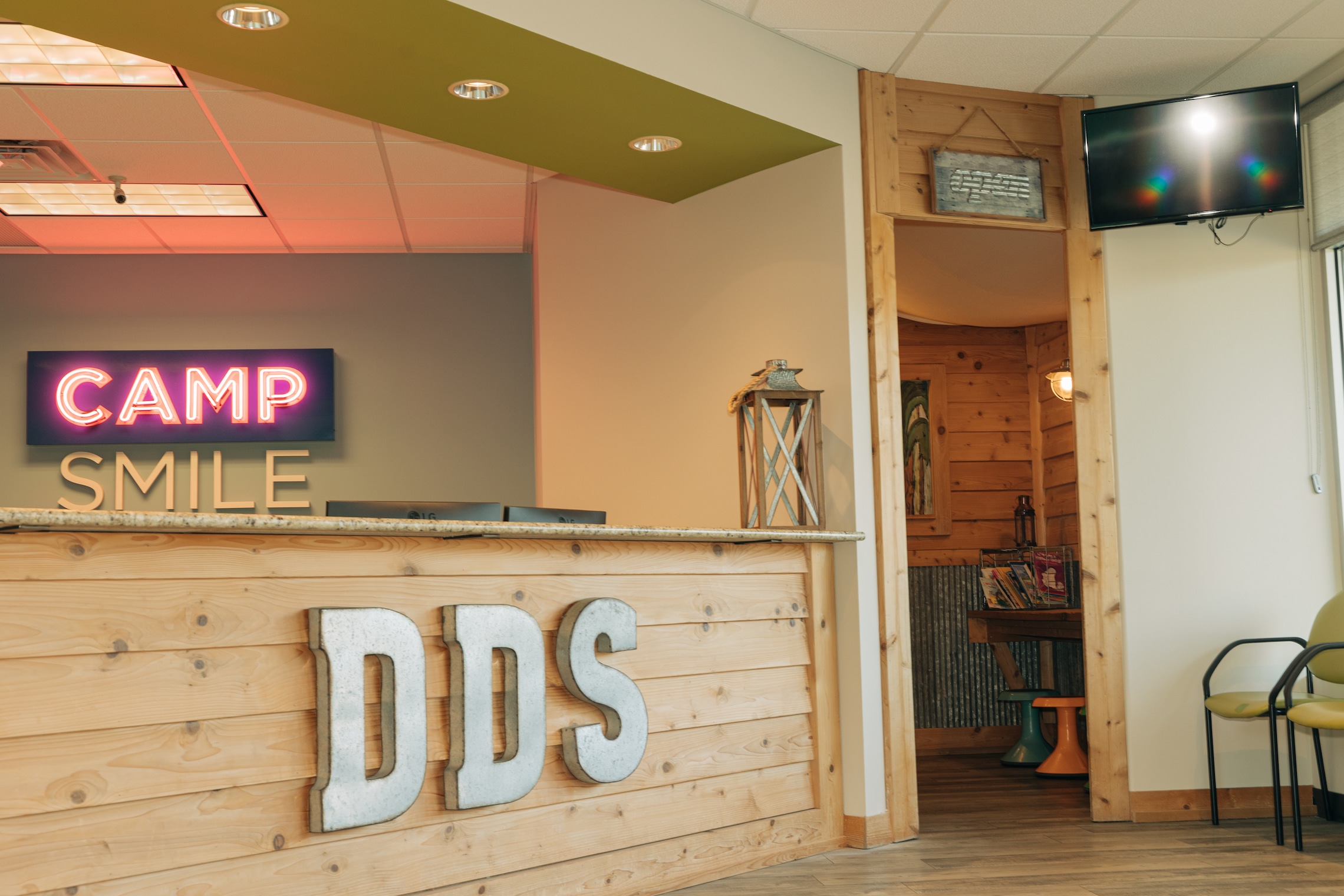 Dental office reception area with wooden decor, neon "Camp Smile" sign, and "DDS" letters. Cozy waiting area with chair and decorative lantern.