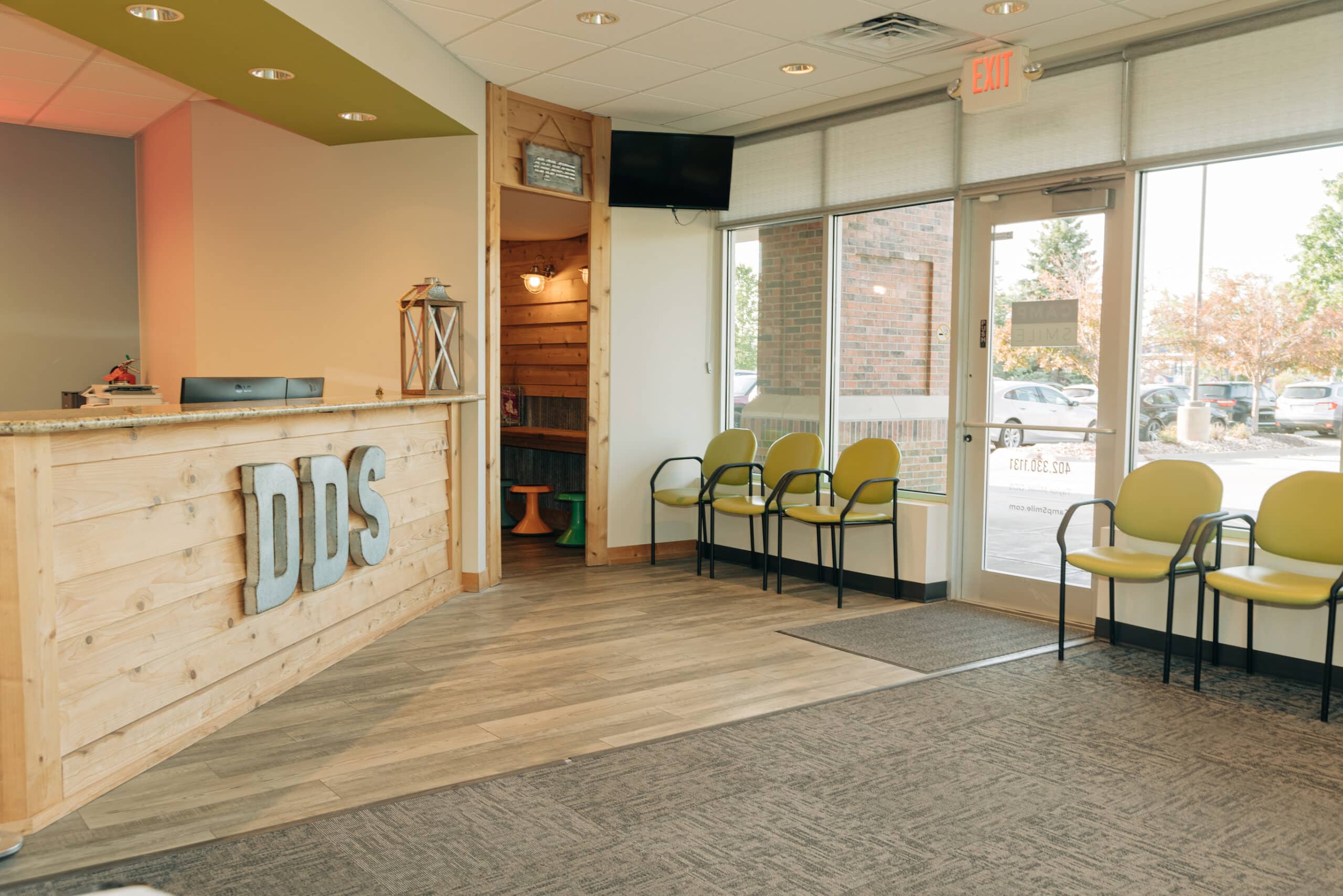 A modern dental office waiting area with a wooden reception desk, labeled "DDS," and several chairs near large windows, overlooking a parking lot.