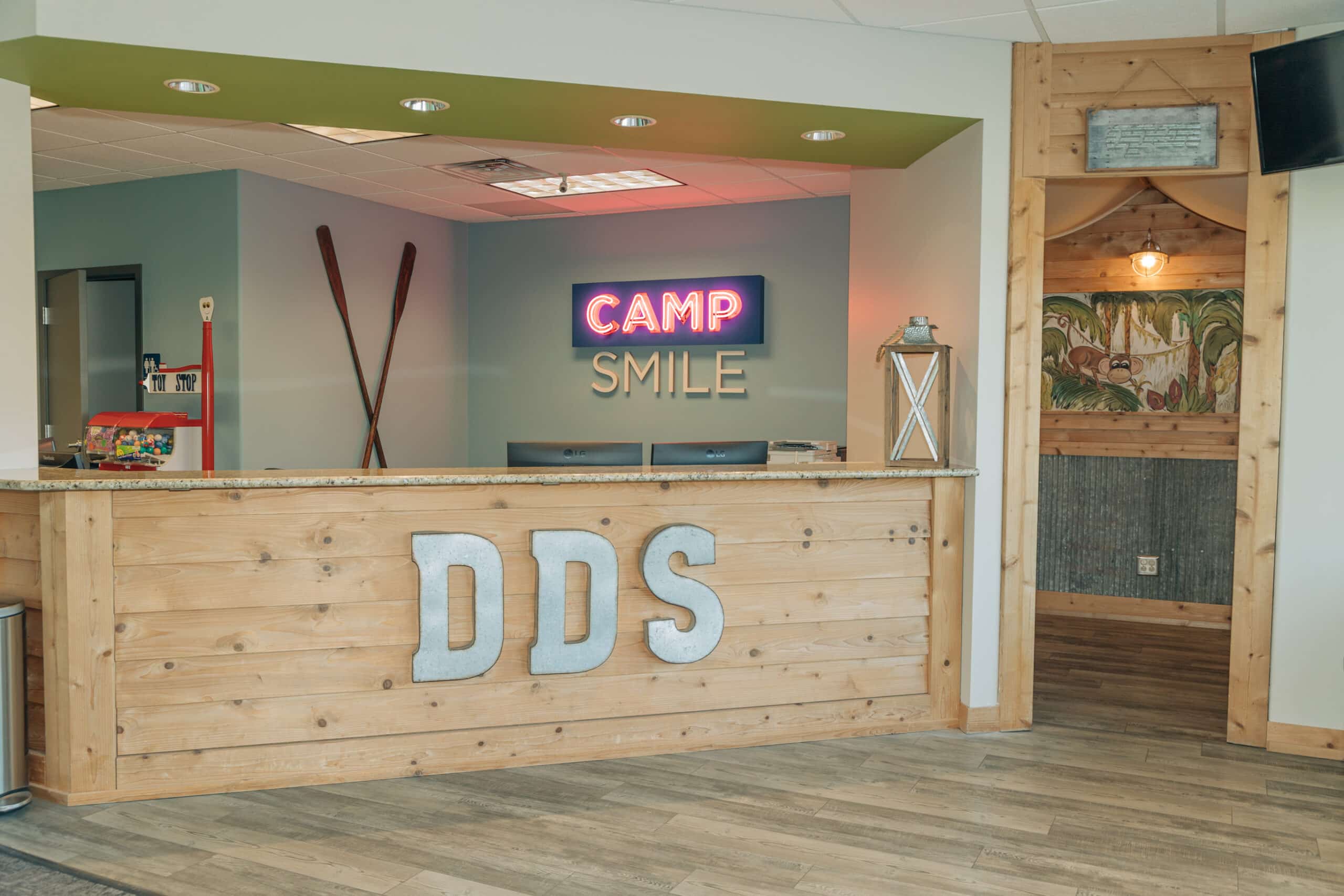 A wooden reception desk with "DDS" letters, and a neon "Camp Smile" sign in an office area, featuring rustic decor.