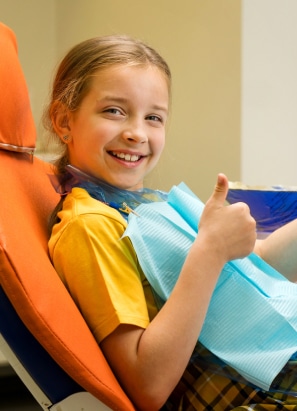 A smiling child sitting in an orange dental chair gives a thumbs-up, wearing a dental bib in a clinic setting.