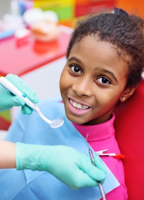 A child is smiling while sitting in a dental chair, receiving a dental check-up from a person wearing green gloves, holding dental tools.