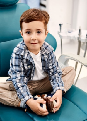 A child sits cross-legged on a dental chair, wearing a plaid shirt and looking at the camera, in a dental office environment.