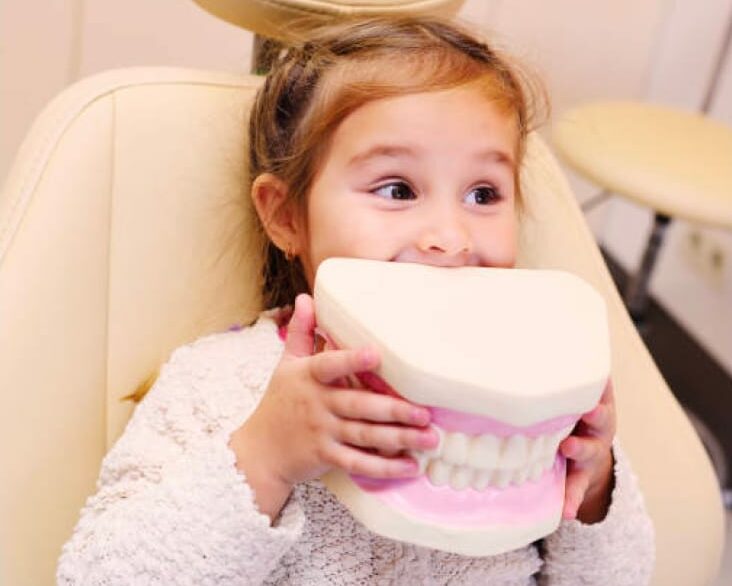 Young girl in dentist chair holding up a giant, plastic replica of teeth.