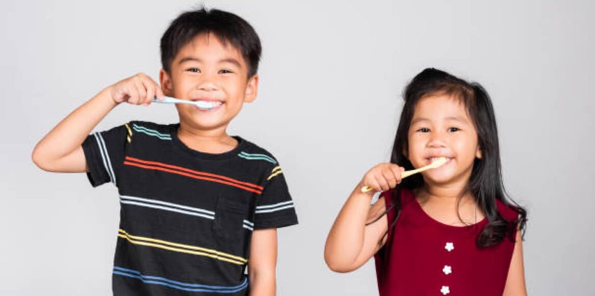 Two young children with brown hair, brushing their teeth in front of a plain gray background.