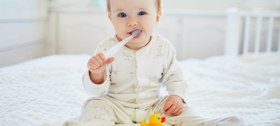 A baby in white pajamas sits on a bed, holding a toothbrush. A yellow rubber duck is placed in front of the baby.