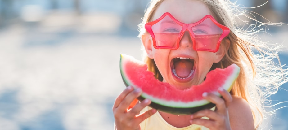 A child, wearing star-shaped sunglasses, joyfully eats a slice of watermelon at the beach, with a blurry background of sand and sunlight.