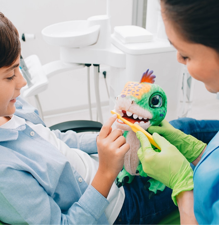 doctor showing boy how to brush teeth using a stuffed dinosaur toy