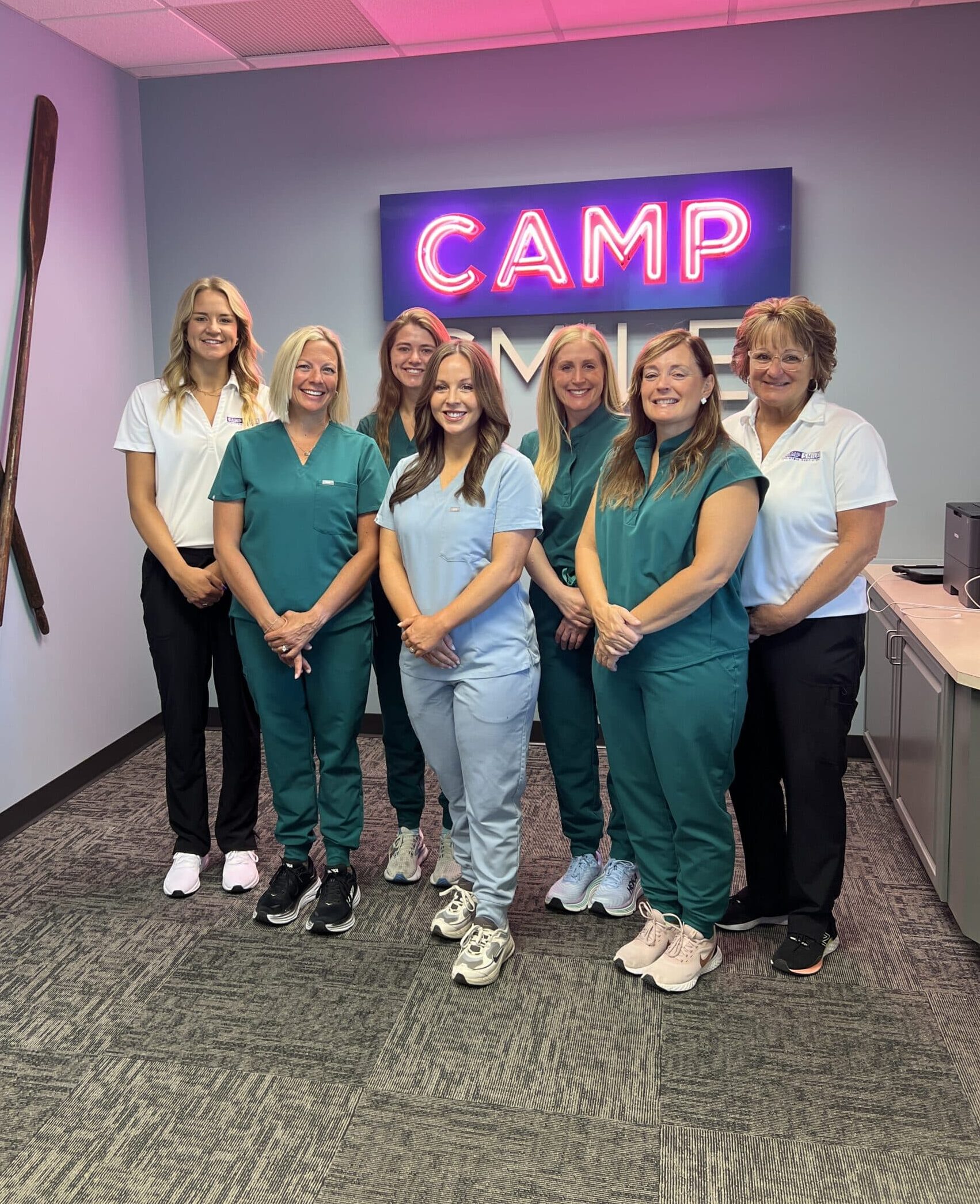 Seven people in dental uniforms stand smiling in front of a neon "CAMP SMILE" sign, inside a room with light blue walls and a ceiling light.