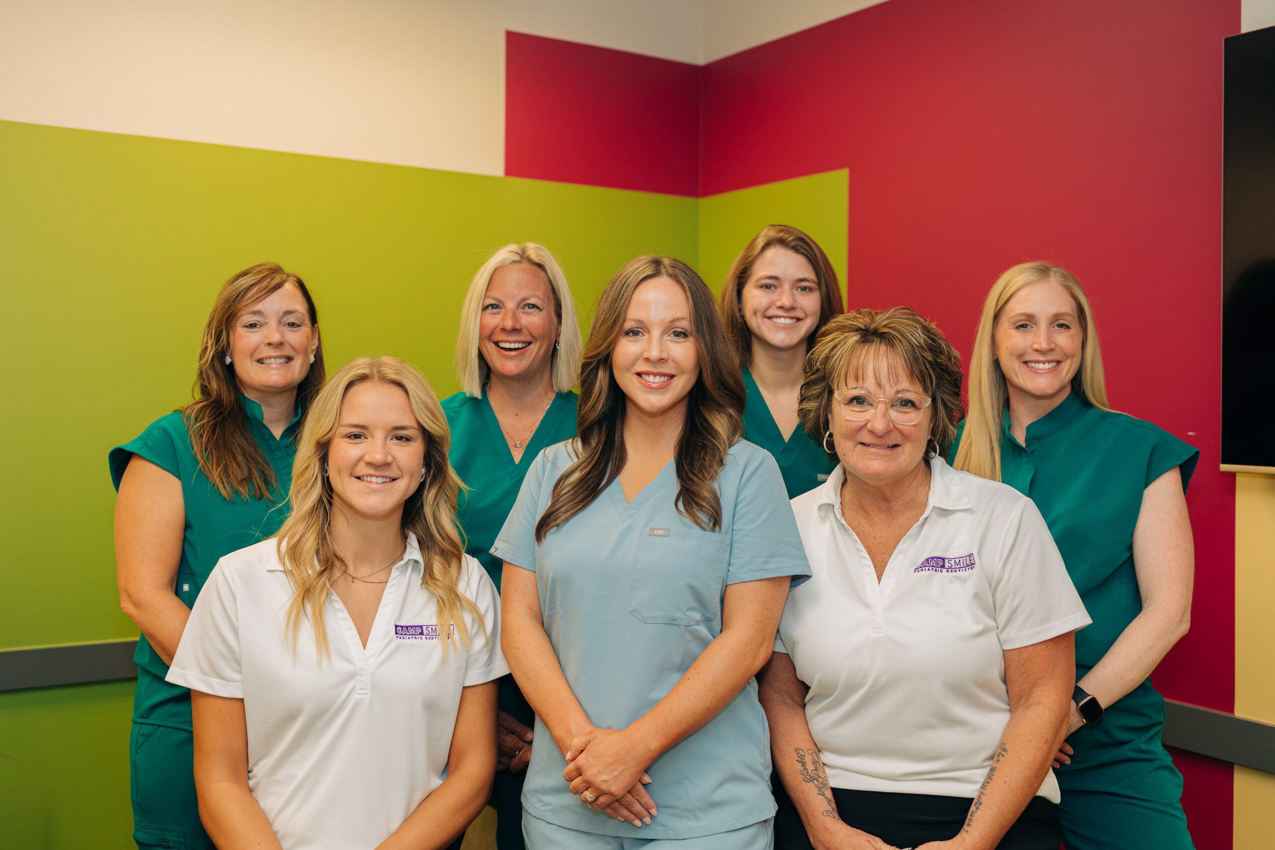 Seven people wearing uniforms smile in a brightly colored room. The group stands close together, suggesting a professional or teamwork-related setting.