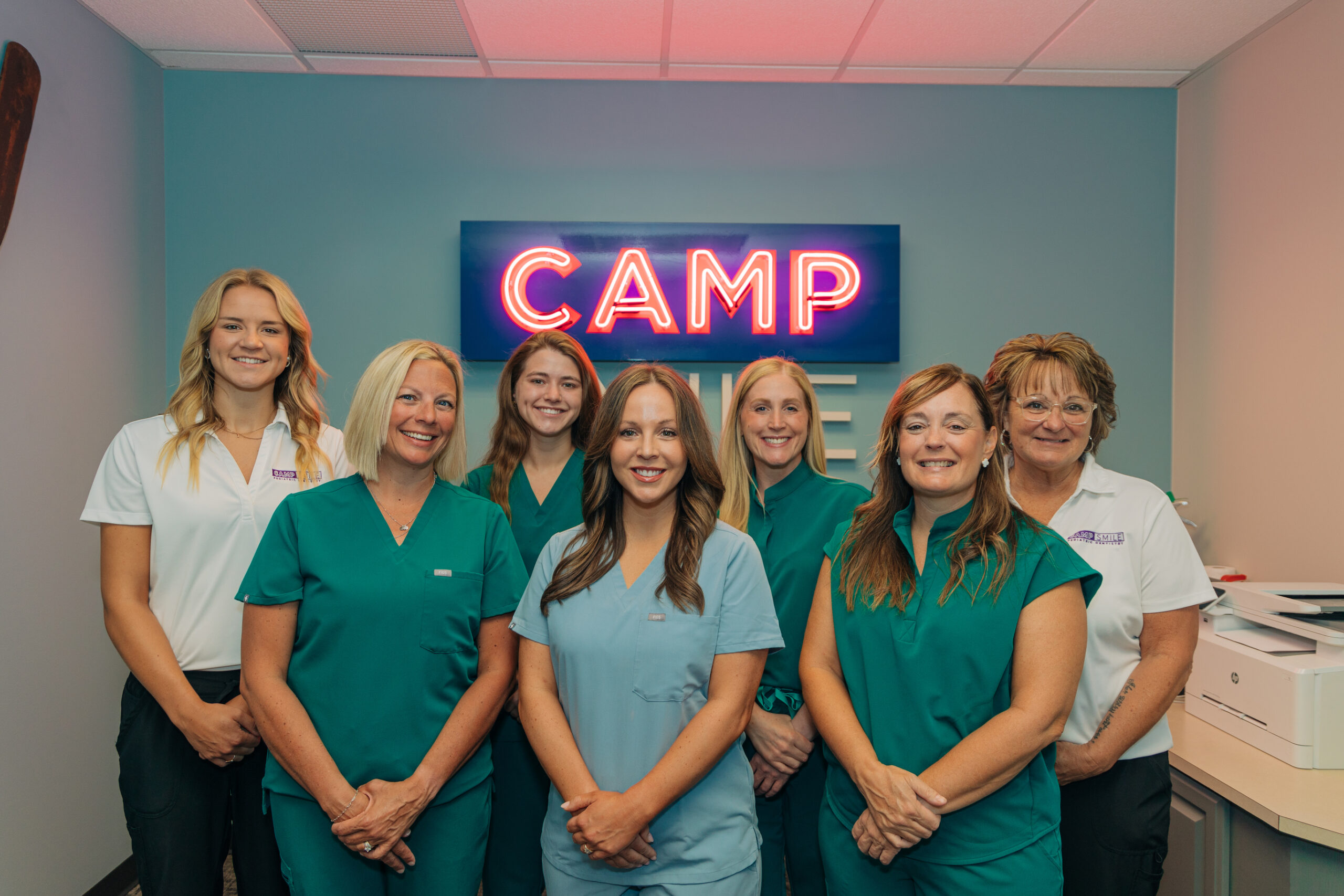 Seven people in uniforms stand smiling in front of a neon "CAMP SMILE" sign, inside a room with light blue walls and a ceiling light.