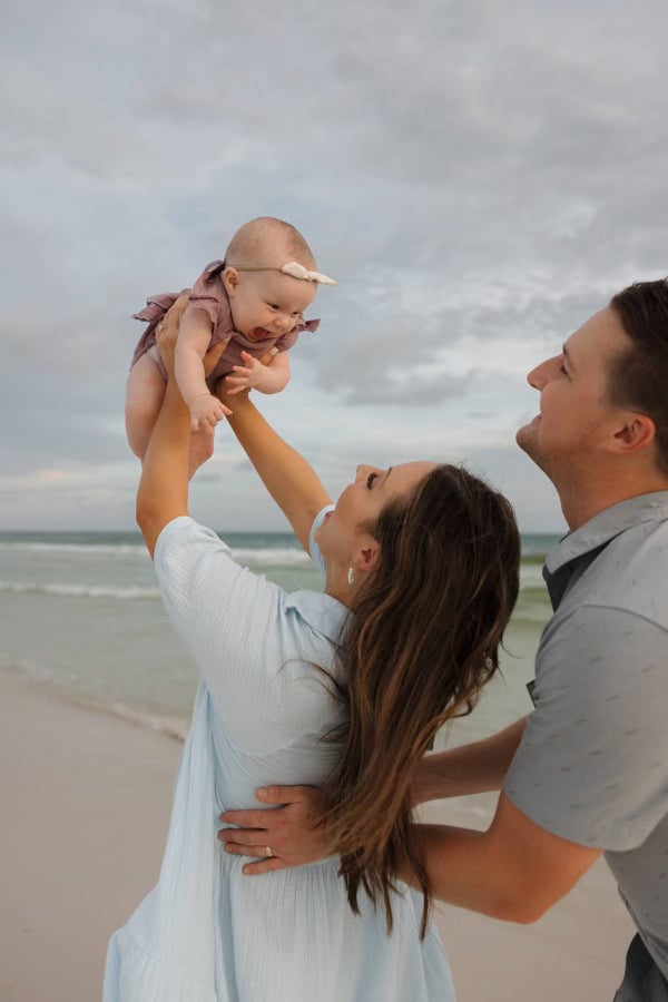 A person lifts a smiling baby on a cloudy beach, while another person stands nearby, creating a joyful family moment by the sea.