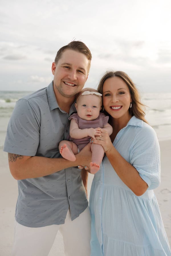 A person holds a baby with another person at a beach. They are smiling, with the ocean and sky in the background.