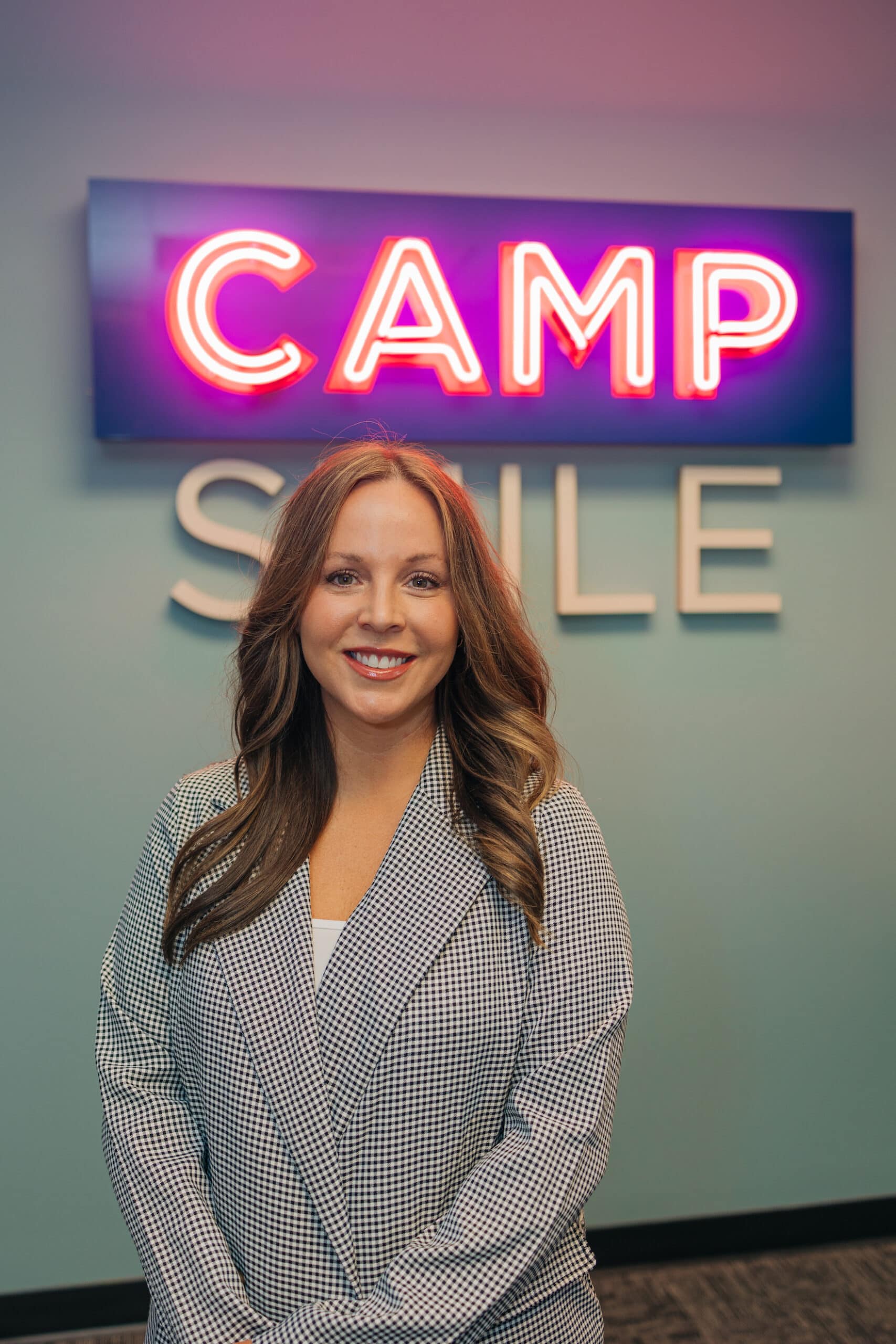 A person smiles in front of a neon sign reading "CAMP SMILE" on a blue wall, exuding a cheerful and welcoming atmosphere.